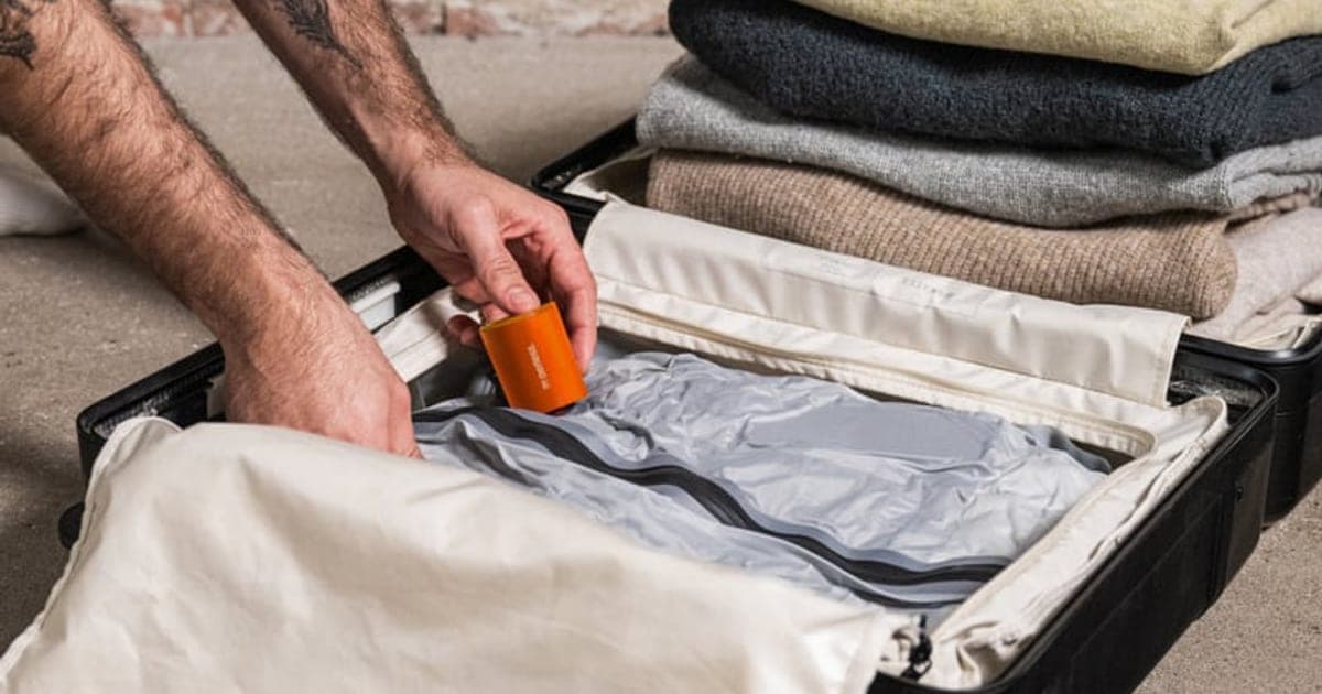 Compressed grey vacuum travel bags lying flat in an open suitcase, with a small orange pump on top and folded sweaters beside them