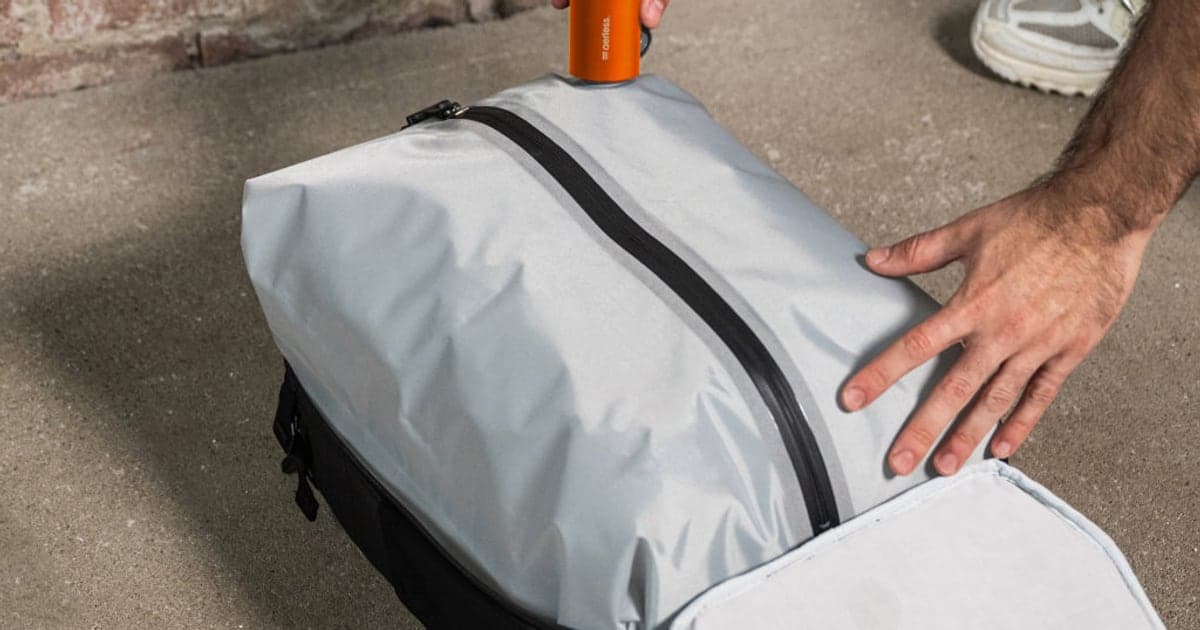 A hand pressing a small orange battery pump onto the valve of a grey vacuum bag packed inside an open backpack